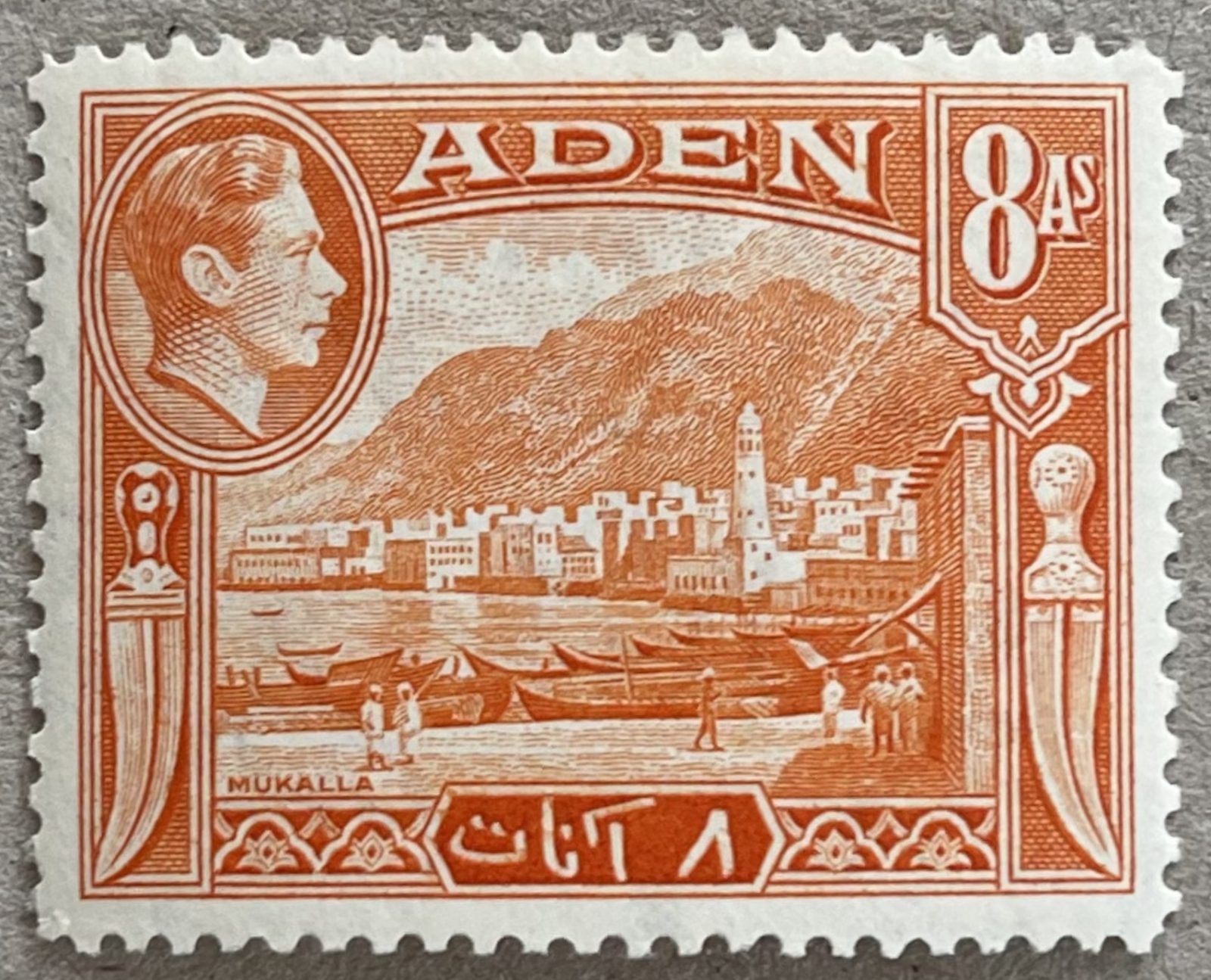 Aden 1939 eight annas stamp in red-orange: George VI portrait, the harbour at Mukalla with dhows on the beach, a tall white tower and mountains behind, curved daggers down each side