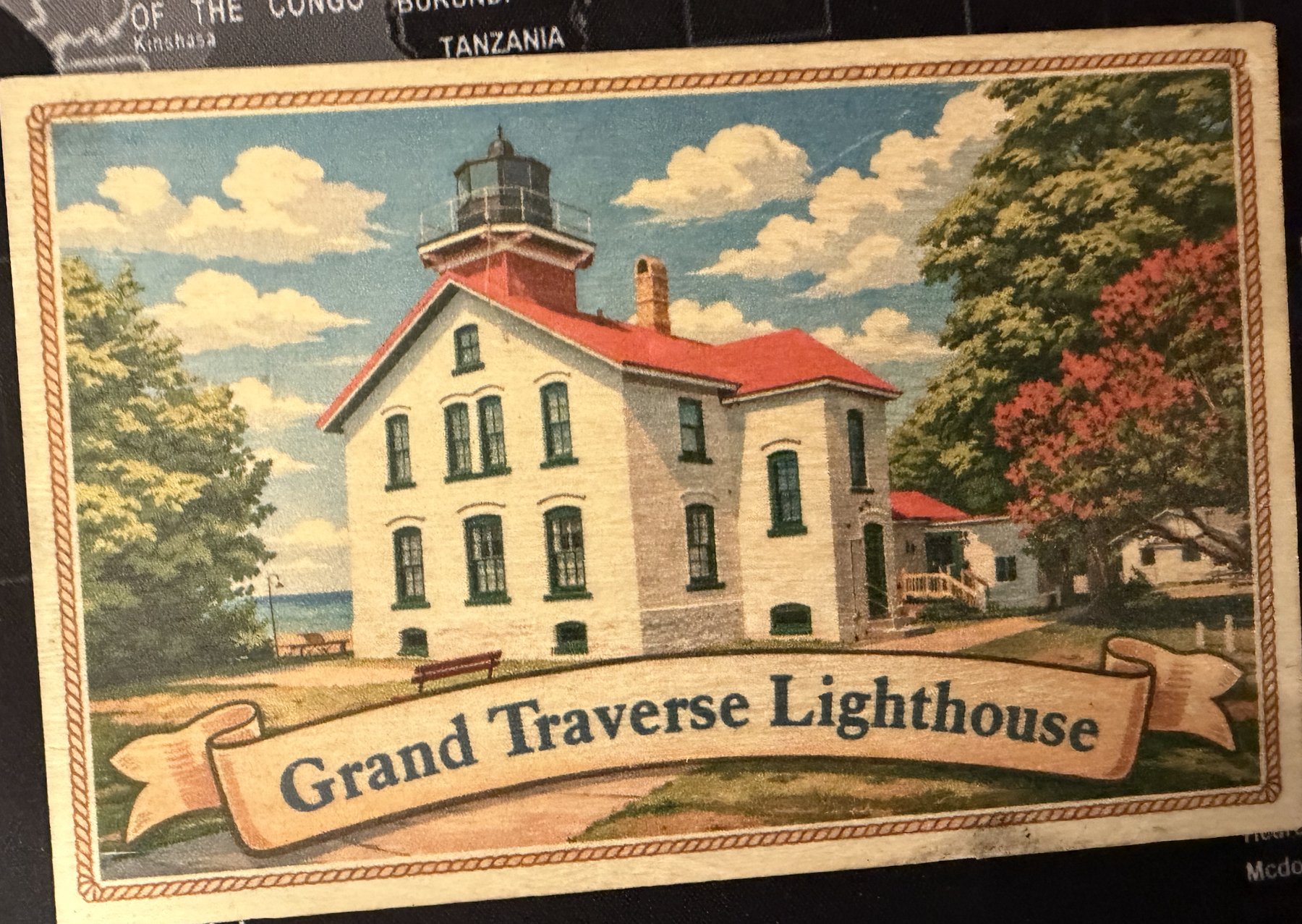 Wooden postcard front: Grand Traverse Lighthouse, a white brick keeper’s house with a red roof and the lantern on top, trees around it and a glimpse of Lake Michigan