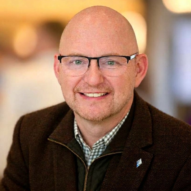 Portrait of NCS smiling, wearing black-framed glasses, a brown tweed jacket and a checked shirt