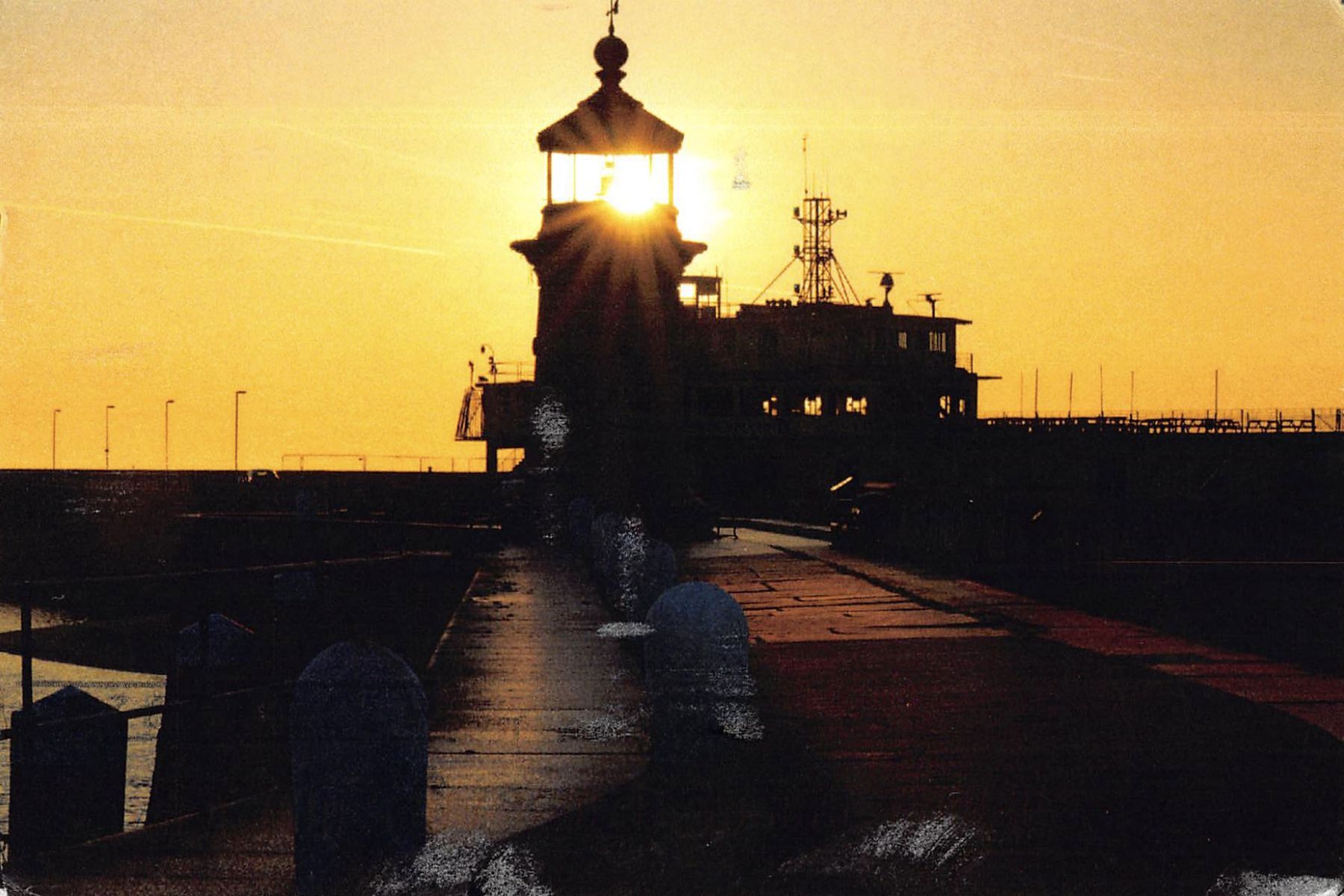 Postcard front: Ramsgate West Pier lighthouse in silhouette at sunrise, the sun glowing inside its lantern, with the pier and bollards in the foreground